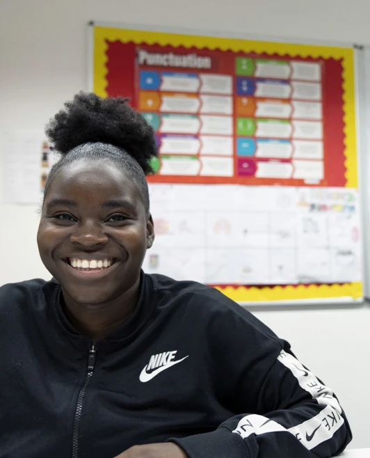 A young female student wearing a black Nike jacket sits at a desk in a classroom, smiling brightly. Behind her, a colorful bulletin board displays educational materials. A young female student wearing a black Nike jacket sits at a desk in a classroom, smiling brightly. Behind her, a colorful bulletin board displays educational materials.